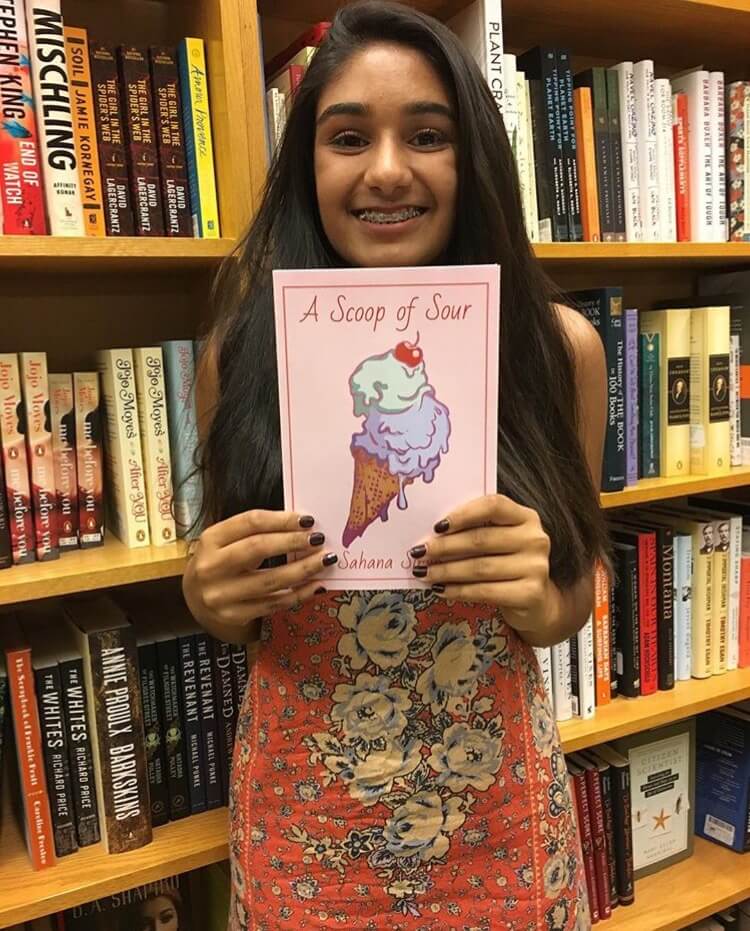 Young author and writing scholarship benefactor, Sahana Singh, standing in front of bookshelves with a copy of her published novel titled A Scoop of Sour.